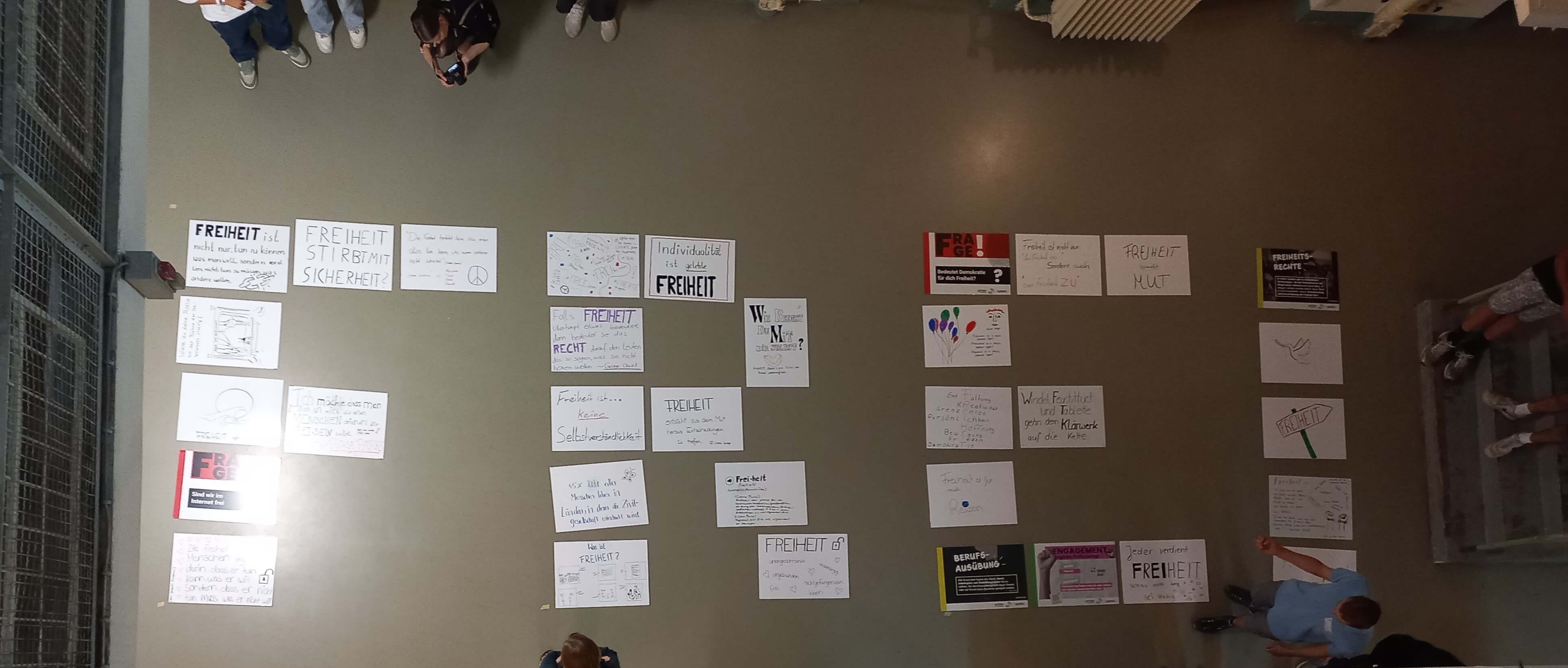 KI generiert: Das Bild zeigt zahlreiche Plakate auf einem Boden ausgelegt, die sich thematisch mit "Freiheit" und ähnlichen Konzepten befassen. Menschen schauen von den Rändern aus auf die Plakate herab.