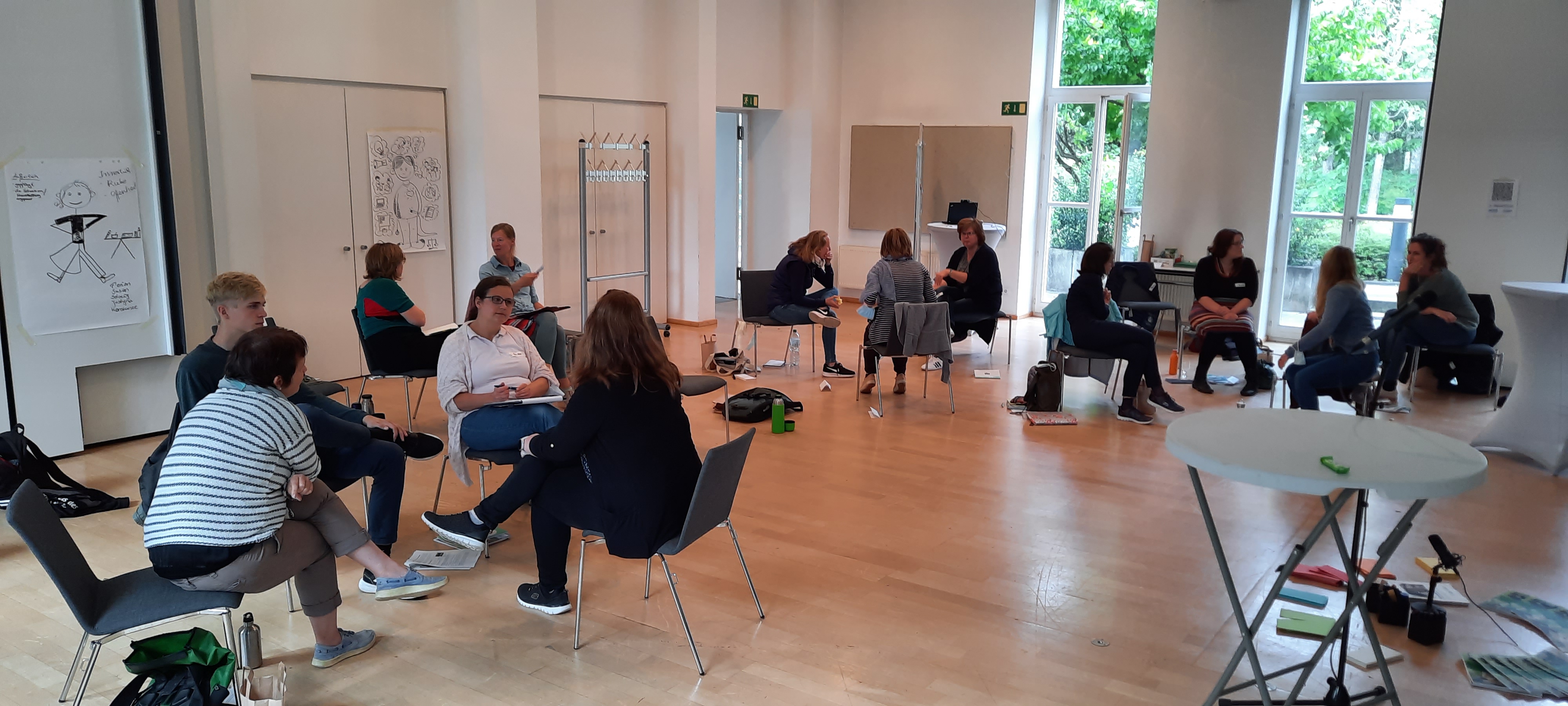 KI generiert: Das Bild zeigt mehrere Menschen, die in einer hellen, geräumigen Umgebung in kleinen Gruppen zusammensitzen und sich unterhalten. Es scheint sich um eine Art Workshop oder Diskussionsrunde zu handeln.