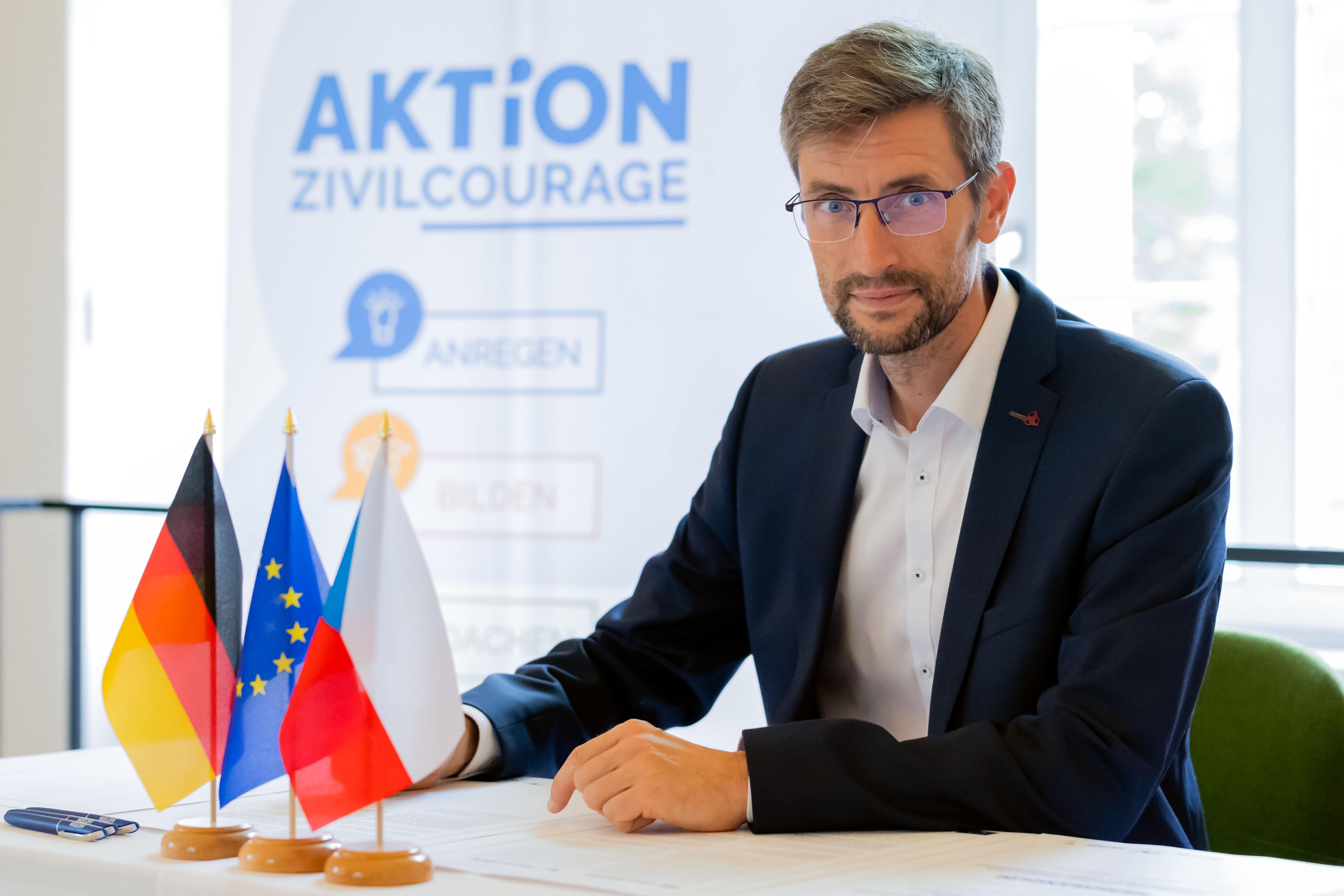 KI generiert: Ein Mann in einem Anzug sitzt an einem Tisch mit mehreren Flaggen (Deutschland, Europäische Union, Tschechien) und einem Banner im Hintergrund mit der Aufschrift „Aktion Zivilcourage“. Er scheint an einem förmlichen Anlass teilzunehmen.