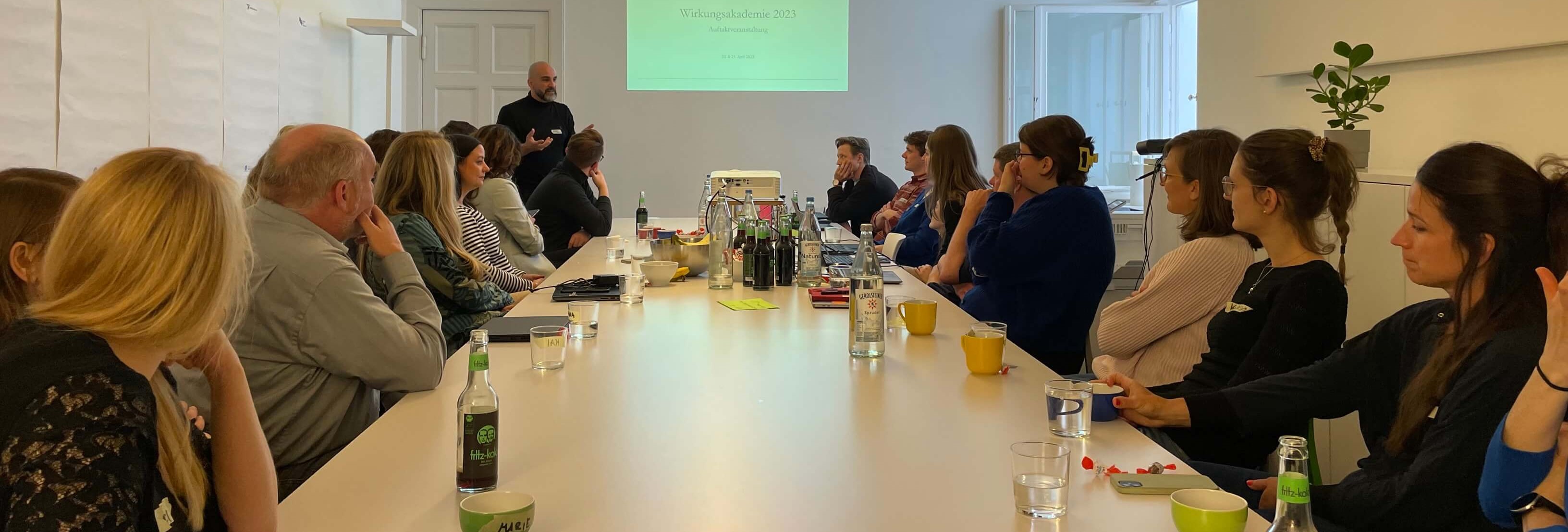 KI generiert: Das Bild zeigt eine Gruppe von Menschen, die in einem Konferenzraum an einem langen Tisch sitzen und anscheinend einem Vortrag oder einer Präsentation folgen. Im Vordergrund stehen Getränke und Schreibutensilien, während eine Person vorne spricht und eine Präsentation projiziert wird.