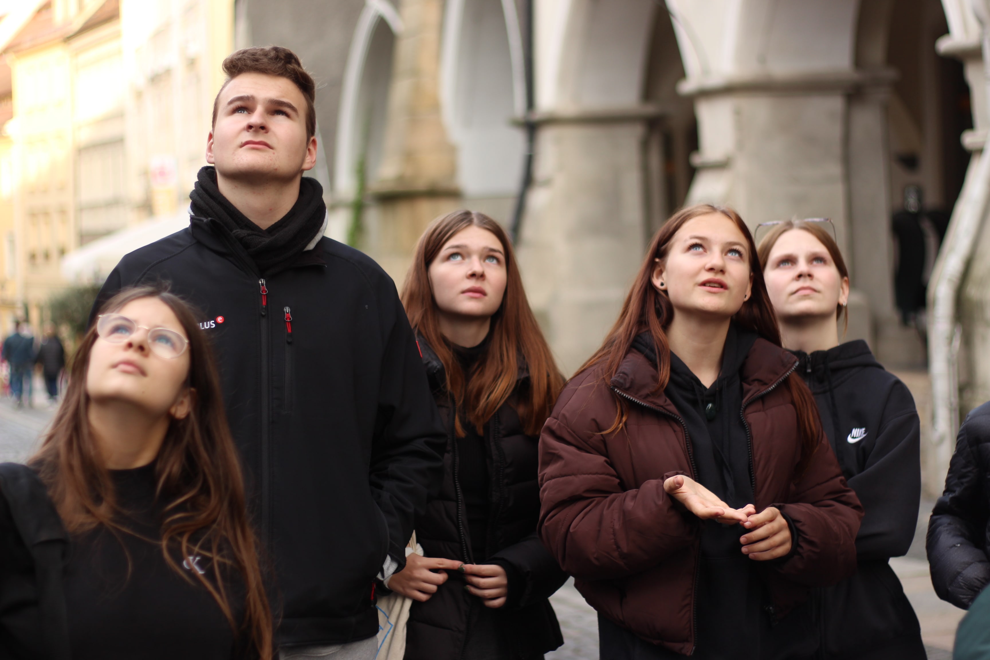 KI generiert: Eine Gruppe von fünf jungen Menschen steht draußen in einer städtischen Umgebung und blickt interessiert nach oben. Im Hintergrund sind Gebäude mit Bögen zu sehen.
