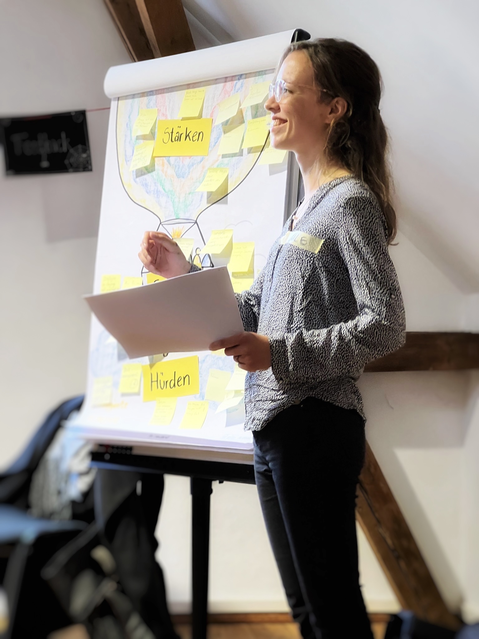 KI generiert: Das Bild zeigt eine Person, die vor einem Flipchart steht und anscheinend eine Präsentation hält. Auf dem Flipchart sind Notizen und die Wörter "Stärken" und "Hürden" zu sehen.